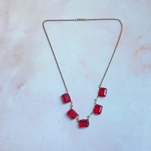 May include: A gold chain necklace with five red gemstone pendants and small white pearl accents.