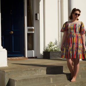 May include: A woman in a colorful, patterned dress stands on a set of stairs in front of a white house with a blue door.