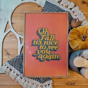 May include: A wooden sign with a red background and black text that reads "Oh Fall it's nice to see you again." The sign is surrounded by fall decor, including a velvet pumpkin, a woven rug, and wooden logs.