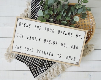 Op de afbeelding: Een houten bord met een witte achtergrond en zwarte tekst die luidt: "Bless the food before us, the family beside us, and the love between us. Amen."