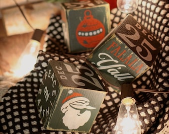 May include: Three green wooden blocks with Christmas-themed designs and text. One block features a Santa Claus illustration, another a red ornament, and the third reads "25 DEC" and "Peace on Earth". A string of lights and a red decorative element are also present.