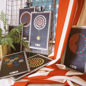 Peut inclure: Cinq impressions encadr&eacute;es noires avec des motifs de feux d'artifice color&eacute;s. Les impressions sont accroch&eacute;es &agrave; un rack en fil de fer avec un tissu ray&eacute; rouge, blanc et bleu drap&eacute; sur le bas. Les impressions portent des num&eacute;ros, dont "No. 79" et "No. 105".