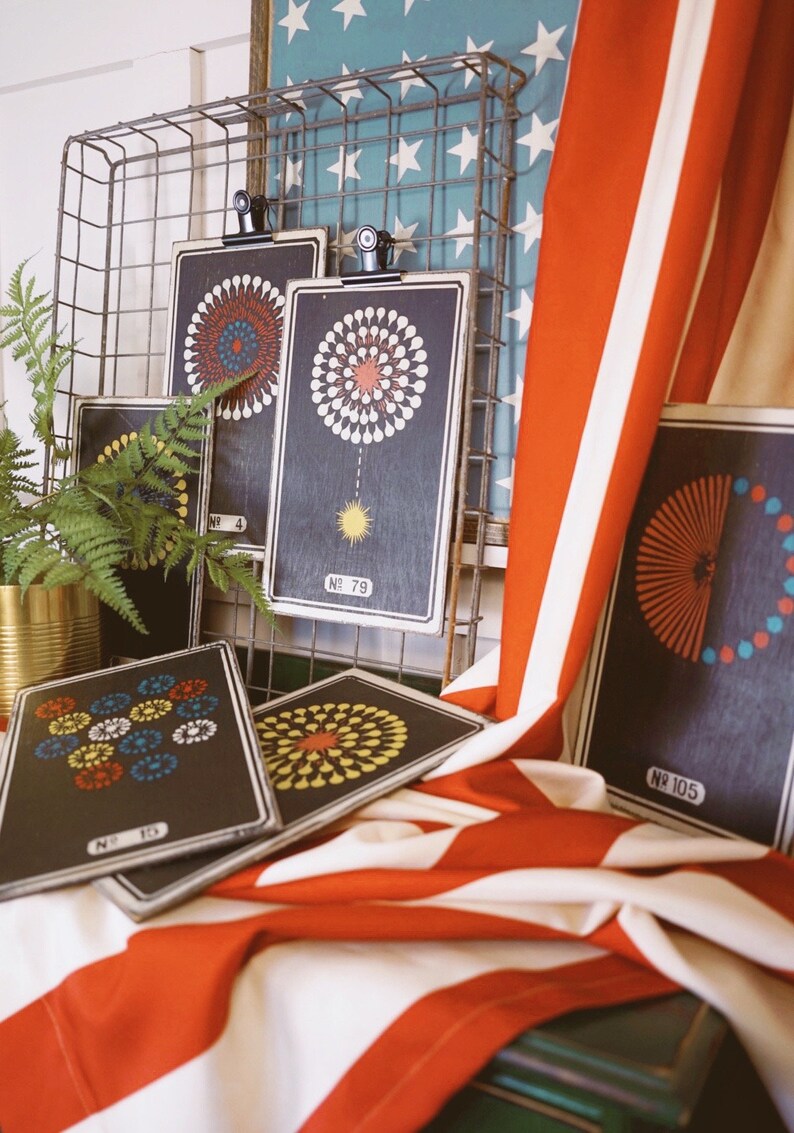 Peut inclure: Cinq impressions encadr&eacute;es noires avec des motifs de feux d'artifice color&eacute;s. Les impressions sont accroch&eacute;es &agrave; un rack en fil de fer avec un tissu ray&eacute; rouge, blanc et bleu drap&eacute; sur le bas. Les impressions portent des num&eacute;ros, dont "No. 79" et "No. 105".