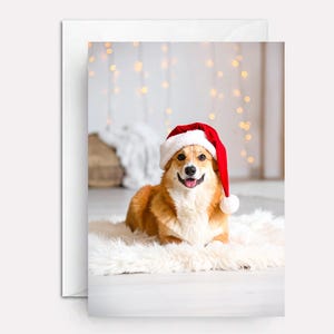 May include: A greeting card featuring a smiling corgi wearing a red and white Santa hat. The dog is lying on a fluffy white rug. The background has soft, blurred lights.