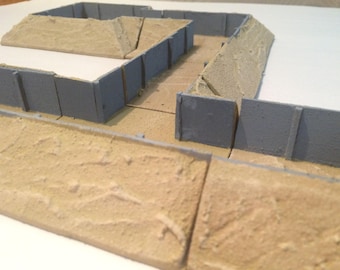 May include: A model of a military bunker with gray walls and sand-colored earthworks. The bunker has a rectangular shape with a small opening in the front.