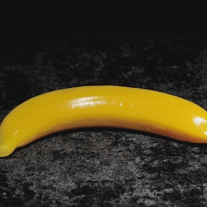 Könnte beinhalten: Ein gelbes, bananenförmiges Objekt mit glänzender Oberfläche. Die Banane ist gebogen und hat eine glatte Textur, mit einer leicht dunkleren Spitze und einem Stiel. Der Hintergrund ist eine dunkle, strukturierte Oberfläche.