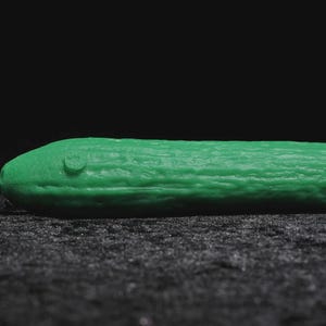 Puede incluir: Un objeto verde brillante con forma de pepino y superficie texturizada. Un pequeño detalle en relieve es visible en un extremo. El fondo es una superficie oscura y texturizada, sobre un fondo negro.