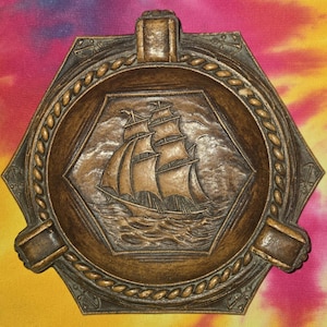 May include: Bronze-colored ashtray with a hexagonal shape. The center features a detailed relief of a sailing ship on the sea. The ashtray has a rope-like border and three built-in rests for cigarettes.