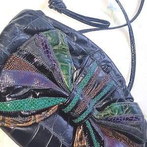 May include: A black leather handbag with a large bow made of different colored leather pieces. The bag has a long strap and a textured surface.