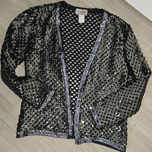 May include: A black sequined cardigan with a mesh panel down the front. The cardigan has a silver trim around the edges and a label that reads "Sherri Hill".