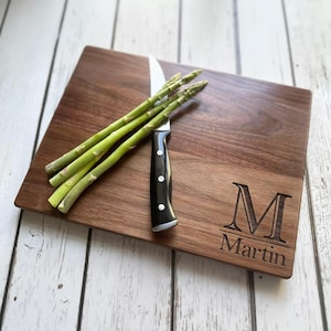 Puede incluir: Tabla de cortar rectangular de madera con un cuchillo y espárragos. La tabla está grabada con la letra "M" y el nombre "Martin". El cuchillo tiene un mango negro con tres remaches plateados. Los espárragos son verdes.