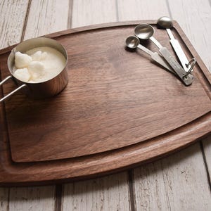 A wooden cutting board with a metal measuring cup filled with sugar, a wooden spoon, and a set of measuring spoons on a white wooden surface.