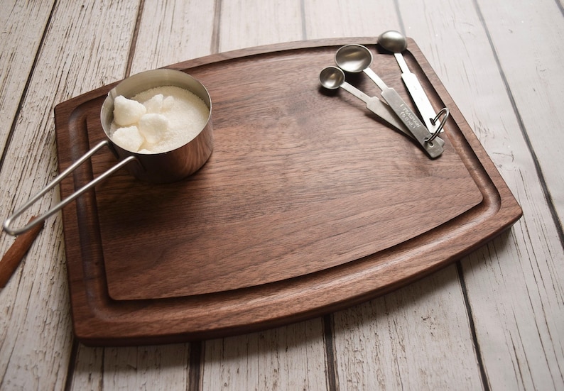 A wooden cutting board with a metal measuring cup filled with sugar, a wooden spoon, and a set of measuring spoons on a white wooden surface.