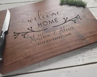 May include: A rectangular wooden chopping board engraved with the words "welcome HOME" and the names "Matthew & Alexis". The address "4246 BAYBERRY LANE" is also engraved. A knife rests on the board's edge, and a sprig of greenery is in the corner.