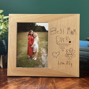 May include: A wooden photo frame with a picture of two people in a field. The frame has a cutout on the right side with the words "Best Mom Ever!" written in black marker. There is a heart and a flower drawn below the text, and the words "Love Ally" are written below the flower.