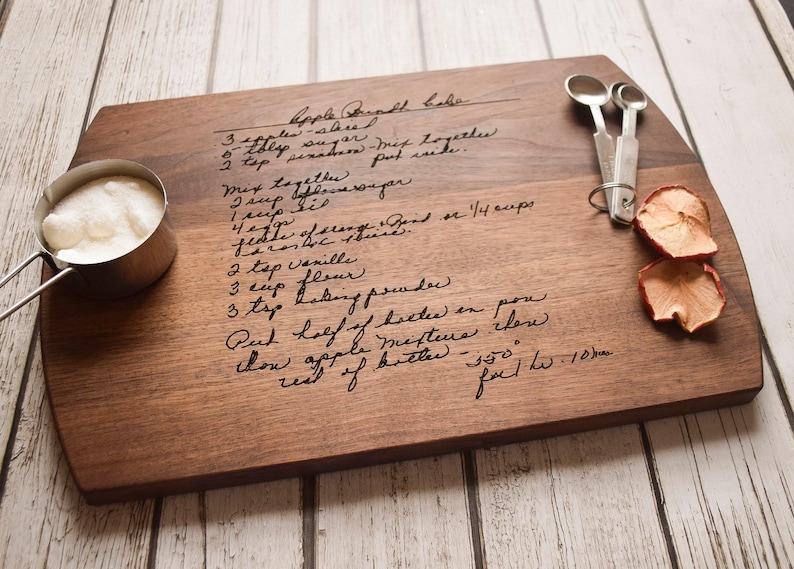 A wooden cutting board with handwritten recipe instructions, a measuring cup, and a pair of scissors, all placed on a white wooden surface.