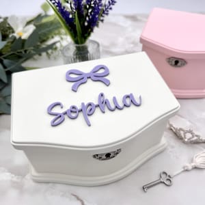 May include: A white jewelry box with a curved design, featuring a purple bow and the name "Sophia". A smaller pink jewelry box is in the background. A silver key and a tiara are also visible.