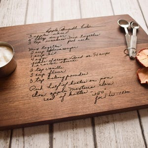 A wooden cutting board with handwritten recipe instructions, a measuring cup, and a pair of scissors, all placed on a white wooden surface.