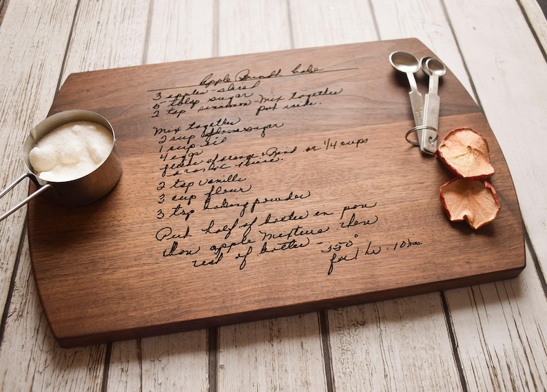 A wooden cutting board with handwritten recipe instructions, a measuring cup, and a pair of scissors, all placed on a white wooden surface.