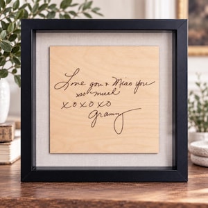 May include: A square black shadow box frame containing a light-colored wooden plaque with dark engraved cursive text that reads: Love you Miao you so much XOXOXO Grammy. The plaque is centered against a light gray fabric backing inside the frame.
