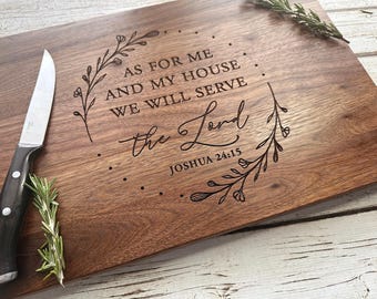 May include: A rectangular wooden cutting board with the engraved text "AS FOR ME AND MY HOUSE WE WILL SERVE the Lord" and "Joshua 24:15". The board has a decorative floral design and is accompanied by a knife and sprigs of rosemary.