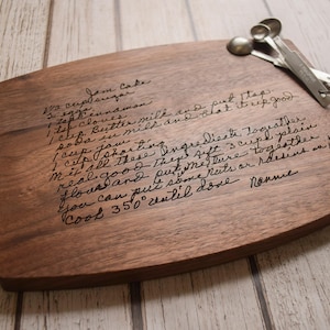 A wooden cutting board with handwritten text and measuring spoons on a white wooden surface.