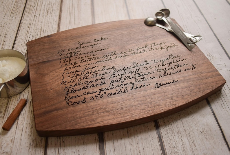 A wooden cutting board with handwritten text and measuring spoons on a white wooden surface.