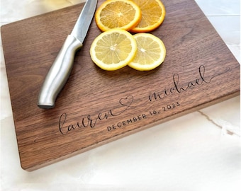 May include: A rectangular wooden cutting board with the names "Lauren & Michael" and the date "December 16, 2023" engraved. The board has a stainless steel knife, lemon and orange slices.