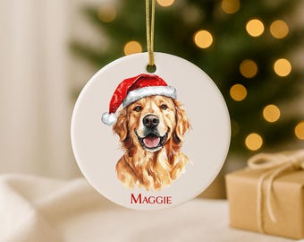 May include: Round Christmas ornament featuring a Golden Retriever wearing a Santa hat. The dog's portrait is detailed with brown and tan fur, a pink tongue, and the name "MAGGIE" in red. The ornament hangs from a gold string.