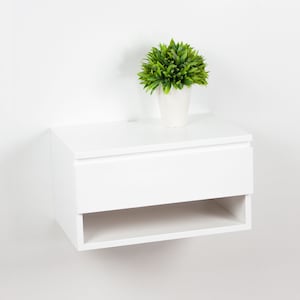May include: White floating nightstand with a single drawer and a shelf. The nightstand is mounted to a white wall and has a small potted plant on top.