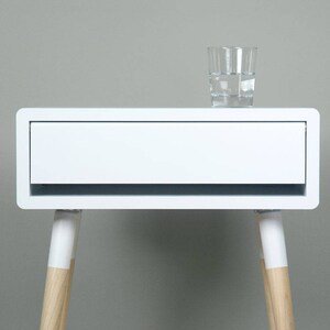 Puede incluir: Mesita de noche blanca con un caj&oacute;n y patas de madera. La mesa tiene un dise&ntilde;o moderno y est&aacute; hecha de laminado blanco.