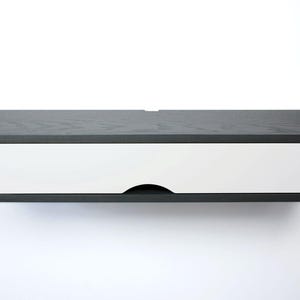 May include: A modern, floating wall-mounted drawer in a minimalist style. The drawer has a dark gray, wood-grain top and sides, with a bright white drawer front. The drawer features a curved handle cutout and is designed to be mounted on a wall.