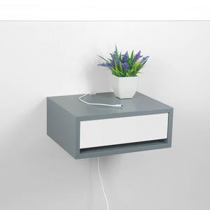 May include: A gray floating nightstand with a white drawer and a white cord running from the back of the drawer. A small potted plant with purple flowers sits on top of the nightstand.