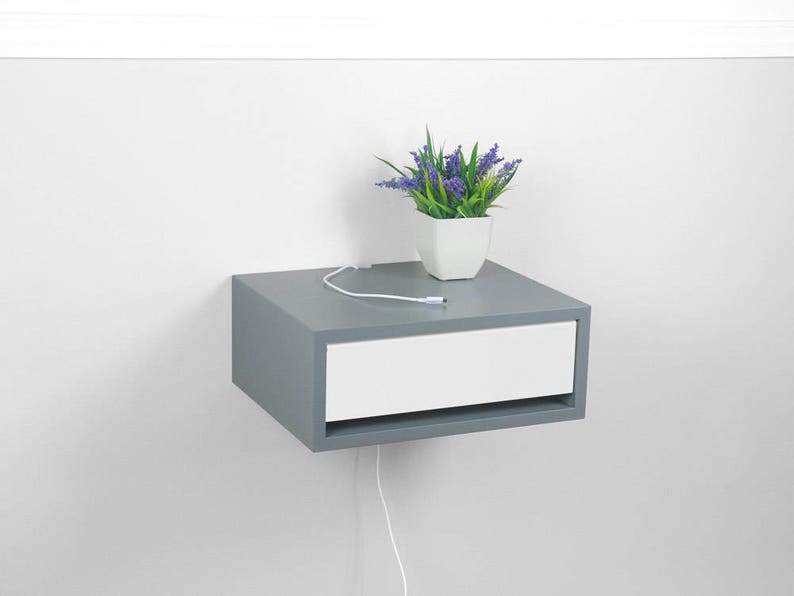 May include: A gray floating nightstand with a white drawer and a white cord running from the back of the drawer. A small potted plant with purple flowers sits on top of the nightstand.