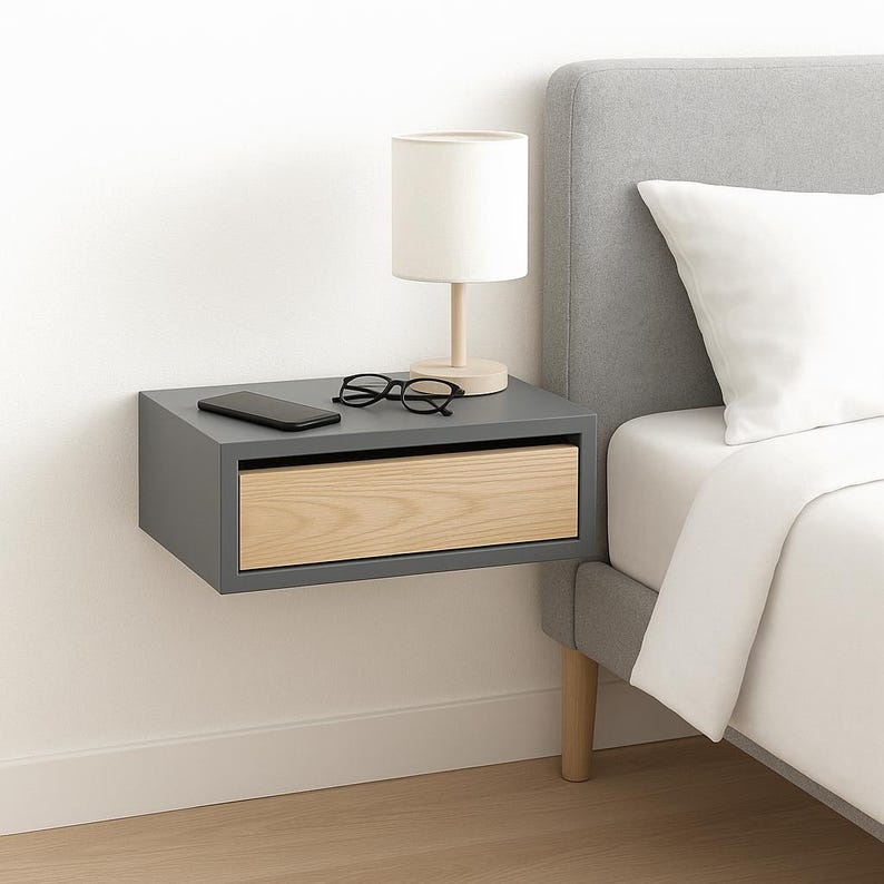May include: A gray floating nightstand with a light wood drawer and a white lamp on top. The nightstand is mounted to a white wall and is next to a gray bed.