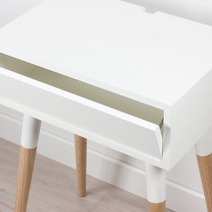 Puede incluir: Mesita de noche blanca con un solo caj&oacute;n y cuatro patas de madera. El caj&oacute;n est&aacute; abierto y la mesa est&aacute; sobre un suelo de madera.