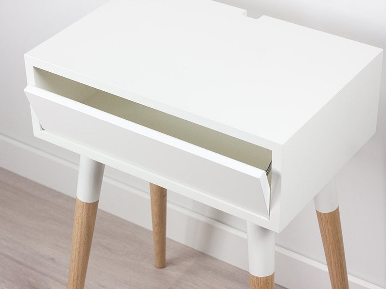 Puede incluir: Mesita de noche blanca con un solo caj&oacute;n y cuatro patas de madera. El caj&oacute;n est&aacute; abierto y la mesa est&aacute; sobre un suelo de madera.