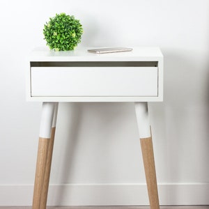 Puede incluir: Mesita de noche blanca con un caj&oacute;n y dos patas de madera pintadas de blanco con un acabado de madera natural en la parte inferior. Una peque&ntilde;a planta verde se encuentra en la parte superior de la mesa.