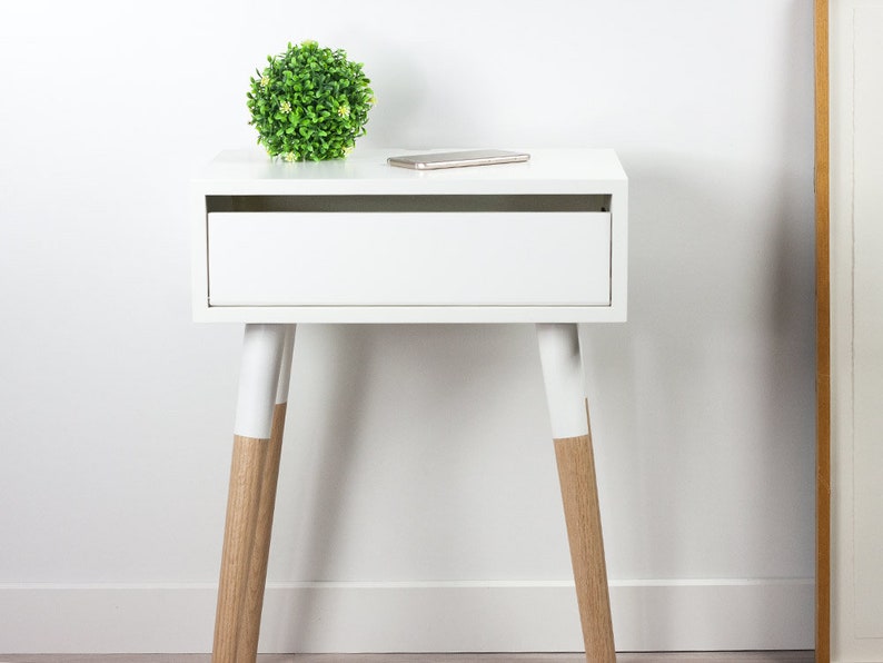 Puede incluir: Mesita de noche blanca con un caj&oacute;n y dos patas de madera pintadas de blanco con un acabado de madera natural en la parte inferior. Una peque&ntilde;a planta verde se encuentra en la parte superior de la mesa.