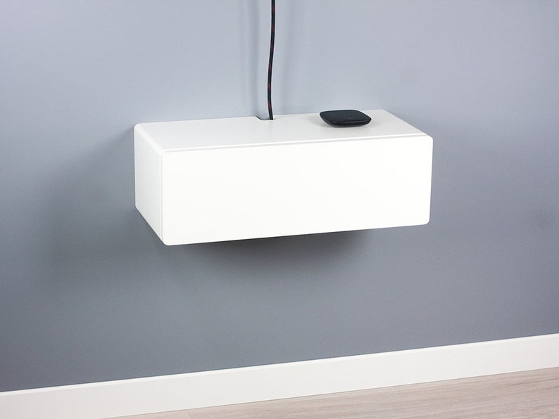 May include: A white floating shelf with a black cord hanging down from the ceiling. A small black device is sitting on the shelf.