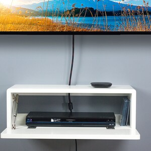 May include: White floating shelf with a black DVD player and a stack of DVDs on the bottom shelf. The top shelf has a small black device on it. The shelf is mounted on a gray wall.