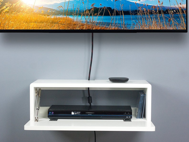 May include: White floating shelf with a black DVD player and a stack of DVDs on the bottom shelf. The top shelf has a small black device on it. The shelf is mounted on a gray wall.