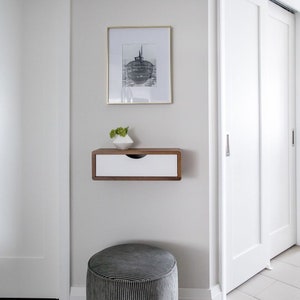 May include: A modern interior scene featuring a framed black and white print above a floating wooden shelf with a white drawer. A small white geometric vase with greenery sits on the shelf. A gray striped ottoman is in the foreground.