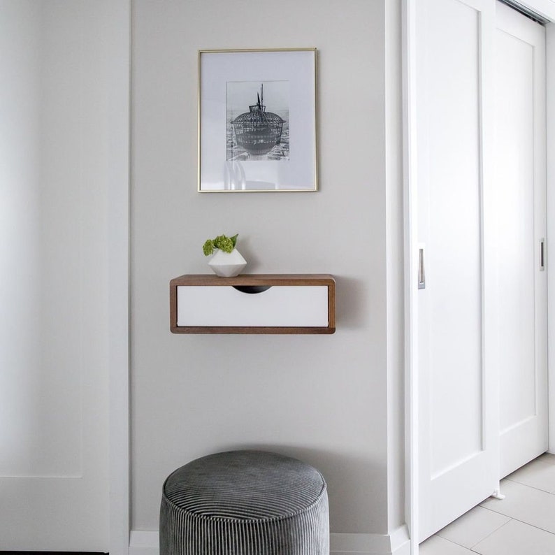 May include: A modern interior scene featuring a framed black and white print above a floating wooden shelf with a white drawer. A small white geometric vase with greenery sits on the shelf. A gray striped ottoman is in the foreground.