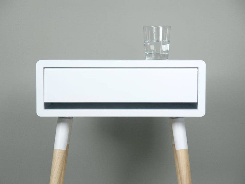 Puede incluir: Mesita de noche blanca con un caj&oacute;n y patas de madera. La mesa tiene un dise&ntilde;o moderno y est&aacute; hecha de laminado blanco.