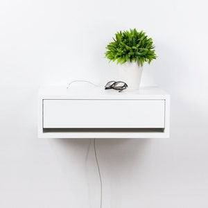 May include: White floating nightstand with a drawer and a small potted plant on top. The nightstand has a white cord hanging from it.