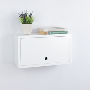May include: White wall-mounted shelf with a single drawer and a small hole in the front. A potted plant and a stack of books are on top of the shelf.