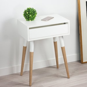Puede incluir: Mesita de noche blanca con un caj&oacute;n y cuatro patas de madera. Las patas est&aacute;n pintadas de blanco con detalles en madera natural. Una peque&ntilde;a planta verde se encuentra en la parte superior de la mesa.