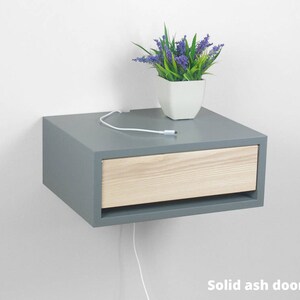 May include: A gray floating nightstand with a single drawer. The drawer front is made of light wood. A small potted plant with purple flowers sits on top of the nightstand.