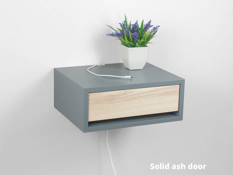May include: A gray floating nightstand with a single drawer. The drawer front is made of light wood. A small potted plant with purple flowers sits on top of the nightstand.